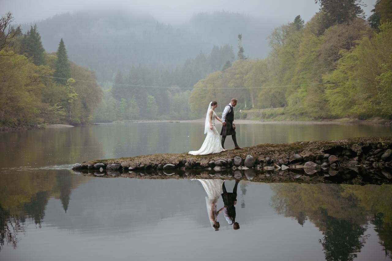 A bride and groom kissing beside a misty Scottish loch, reflected in the water